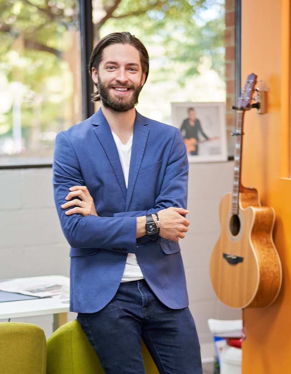 Brian Larsen in a blue blazer, arms crossed and smiling, standing in his office beside an acoustic guitar hanging on an orange wall