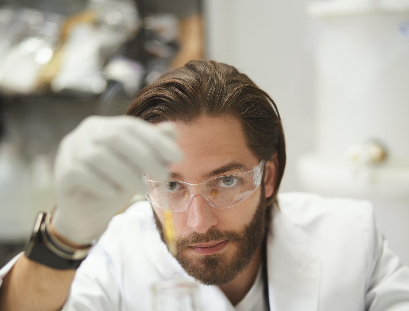 Brian Larsen in a lab coat and safety glasses, holding a pipette in a gloved hand and studying the sample closely