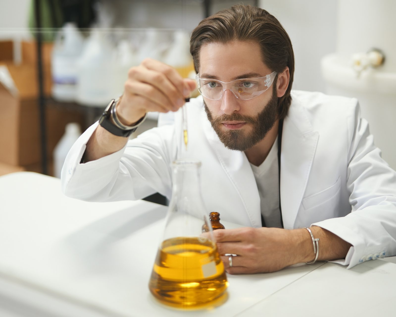 Brian Larsen in a white lab coat and safety glasses, drawing amber liquid from a dropper into an Erlenmeyer flask at a laboratory counter